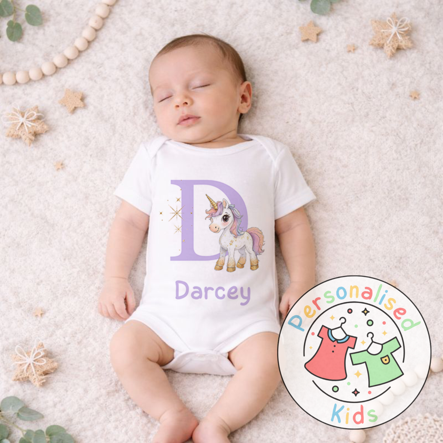 Baby wearing a white onesie with a unicorn design and 'Darcey' text, lying on a textured surface with decorative elements.