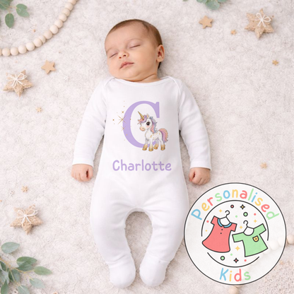 Baby wearing a white onesie with a unicorn design and 'Charlotte' text, lying on a soft surface with decorative elements.